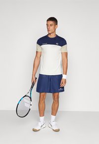 Tennis outfit featuring a cream and navy short-sleeve shirt with blue accents, paired with navy shorts and white athletic shoes.