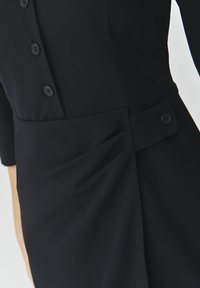 Schwarzes Kleid mit Dreiviertelärmeln, mit einer durchgehenden Knopfleiste, Falten-Details und einer Seitentasche, die mit einem passenden Knopf verziert ist.