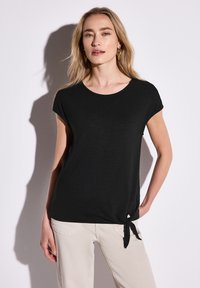 Top nero a maniche corte con scollatura tonda e dettaglio con nodo sul lato, realizzato in tessuto morbido, abbinato a pantaloni di colore chiaro.