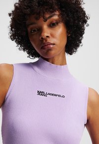 Bele protinė be rankovių palaidinė su aukštu kaklu ir siuvinėtu "KARL LAGERFELD JEANS" logotipu, pasižyminti lygiu audiniu ir prigludusia forma.