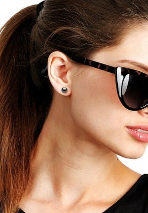 Femme aux cheveux bruns en queue de cheval, portant des boucles d'oreilles clous noires et des lunettes de soleil écaille, regardant vers la droite avec une expression neutre.