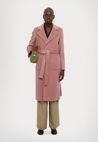 Rosa ullkappa med belagd midja, slagkrage och sidofickor, matchad med beige vida byxor och svarta skor, bärande på en grön väska.