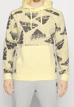Mężczyzna ubrany w żółtą bluzę z kapturem Adidas z czarnymi wzorami logo i szare spodnie dresowe Adidas, stojący na jednolitym tle.