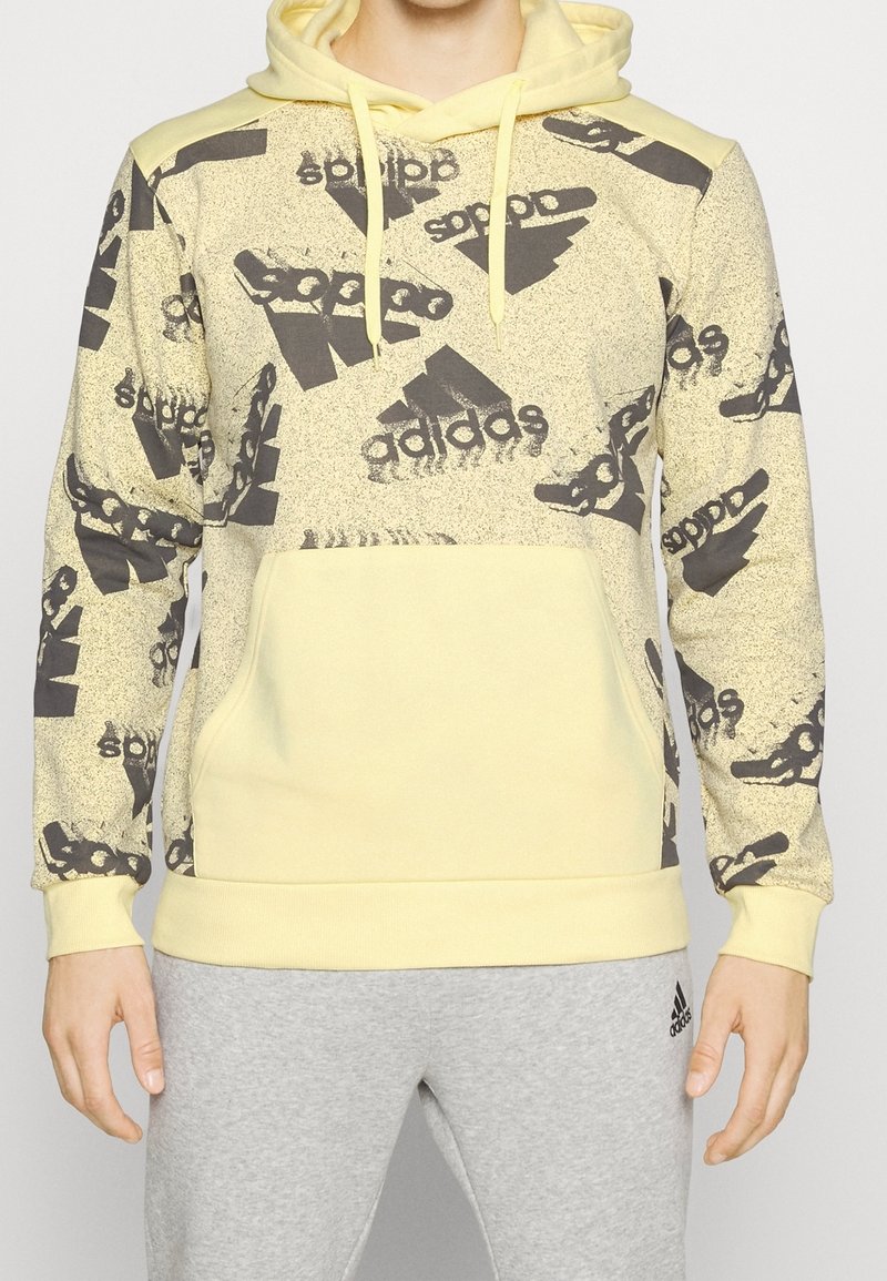 Homme portant un sweat à capuche Adidas jaune avec des motifs de logo noirs et un pantalon de survêtement Adidas gris, debout devant un fond uni.