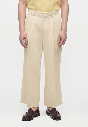 Personne portant un pantalon beige à jambes larges, une chemise jaune rentrée et des mocassins marron foncé, debout sur un fond clair.