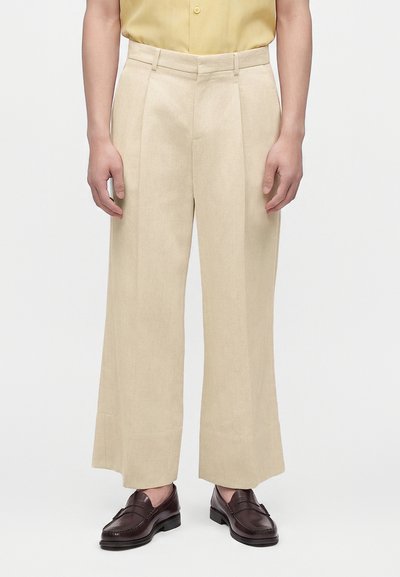 Personne portant un pantalon beige à jambes larges, une chemise jaune rentrée et des mocassins marron foncé, debout sur un fond clair.