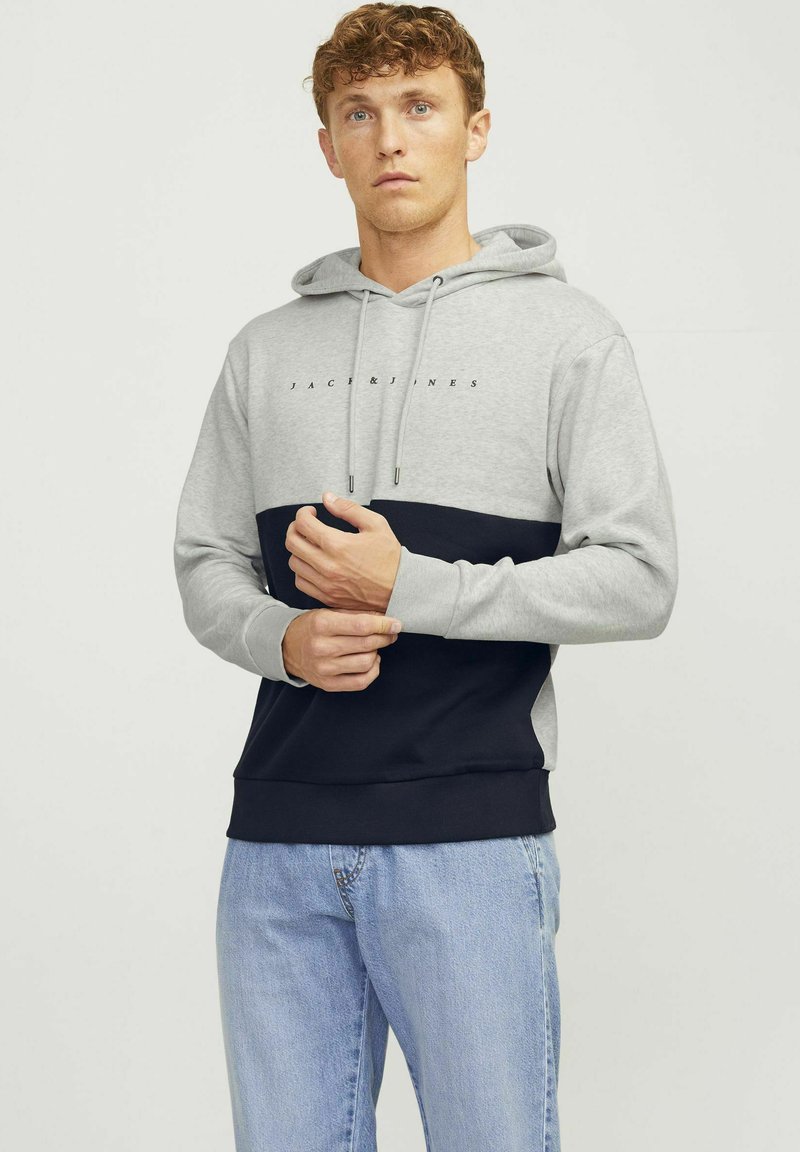 Sudadera color bloque gris y marino con cordón, fabricada en suave algodón, puños acanalados y logo en el pecho; combinada con jeans de color azul claro.