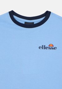 Svetlomodré bavlnené tričko s tmavomodrým rebrovaným golierom a čiernym logom "ellesse", ktoré obsahuje polkruhový slnečný emblém v oranžovej a červenej farbe.