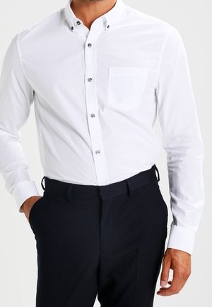 Mand iført en tætsiddende hvid skjorte med brystlomme og sorte bukser, hånd i lommen på en ensfarvet baggrund.