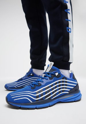 Blaue und schwarze Sportsneaker mit weißen wellenförmigen Streifen, getragen zu schwarzen Hosen und weißen Socken vor hellem Hintergrund.