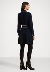Vestido azul-marinho com mangas longas, detalhes brancos ao longo da gola e da cintura, saia evasê, acompanhado de botas pretas até o tornozelo com detalhes em dourado.