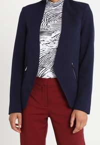 Blazer azul marino con cuello chal, dos bolsillos con cremallera, llevado sobre una blusa blanca con estampado negro y pantalones burdeos. Textura de tela suave.