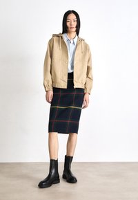 Chaqueta beige con capucha y botones a presión, camisa azul de botón, falda de cuadros azul marino con acentos verdes y rojos, y botas negras hasta el tobillo.