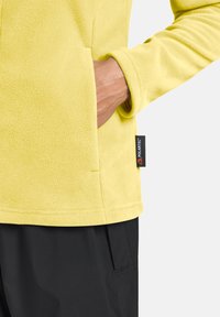 Giacca in fleece giallo brillante con una texture morbida, dotata di una tasca laterale e etichetta Polartec. Abbinata a pantaloni neri.