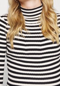 Maglione a collo alto a righe bianche e nere con texture a costine, design aderente e collo alto, che mostra righe orizzontali alternanti.