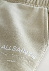 Béžová tkanina s bielym textom "ALLSAINTS" a menším textom v japončine a nápisom "LONDON" vytlačeným nižšie na zloženom oblečení.