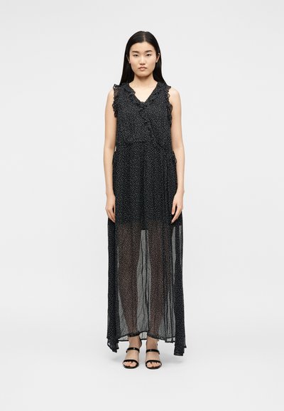 Γυναίκα στέκεται φορώντας μαύρο αμάνικο μάξι φόρεμα με λευκές πουά, διάφανο στρώμα φούστας και μαύρα πέδιλα ανοιχτά στην άσπρη πίσω όψη.