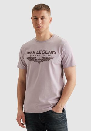 Mężczyzna w jasnofioletowej koszulce PME Legend z ciemnym emblematem w kształcie skrzydeł i napisem, stojący z jedną ręką w kieszeni na jednolitym tle.