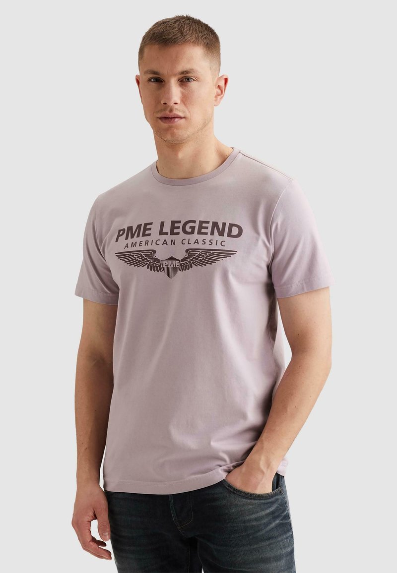 Άνδρας που φορεί ανοιχτό μωβ μπλουζάκι PME Legend με σκούρο έμβλημα φτερού και κείμενο, στέκεται με το ένα χέρι στην τσέπη μπροστά σε απλό φόντο.
