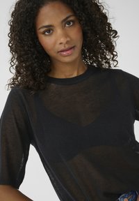 Pull en maille noire transparente à manches courtes, col rond et motif rayé subtil qui crée un effet superposé.
