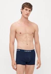Joven con cabello corto que lleva bóxer azul marino de Lacoste, de pie contra un fondo claro, mirando hacia adelante.