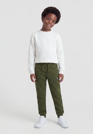 Enfant aux cheveux bouclés portant un sweat blanc, un jogging vert olive et des baskets blanches, debout devant un fond clair uni.