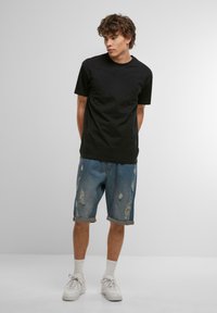 T-shirt en coton noir, coupe ample. Porté avec des shorts en denim déchirés, bleu clair, longueur genou avec ourlets roulés. Baskets blanches et chaussettes.