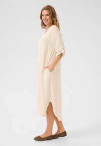 Beige knälång klänning med trekvartsärmar, sidfickor och hög-l�åg fåll. Mjuk textur och avslappnad passform. Bärs med platta bruna skor.