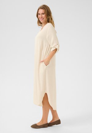 Beige knälång klänning med trekvartsärmar, sidfickor och hög-låg fåll. Mjuk textur och avslappnad passform. Bärs med platta bruna skor.