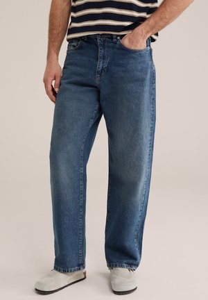 Férfi egyenes szárú kék farmerben, csíkos ingben és világosszürke cipőben, kezét zsebében tartva áll, egyszínű háttér előtt.