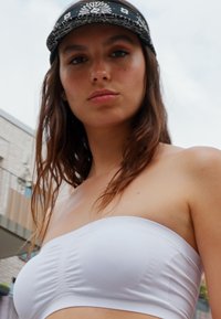 Haut de bikini blanc sans bretelles avec des détails froncés, associé à une casquette noire à motifs. L'arrière-plan présente un bâtiment léger et texturé.
