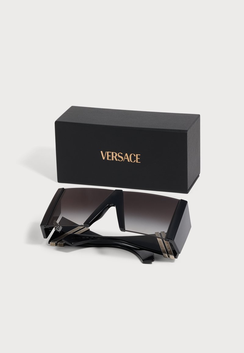 Lunettes de soleil noires surdimensionnées de style visière avec des accents métalliques à côté d'une boîte noire avec le logo Versace doré sur fond blanc.