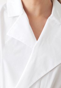 Prenda de color blanco con cuello tipo polo que presenta un diseño de escote en v, textura suave y detalles de tela en capas, enfatizando la sencillez y la elegancia.