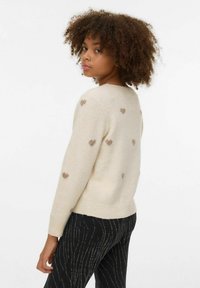 Béžový svetr s malými hnědými srdíčkovými vzory, žebrovaným lemem a manžetami. Modelka má na sobě vzorované černé kalhoty, je otočená zády ke kameře.