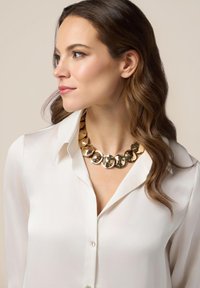 Collana dichiarativa dorata con dischi circolari riflettenti su un modello che indossa una blusa bianca; texture liscia e design audace.