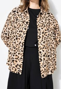 Leopard-print button-up shirt with a collar, short sleeves, and two front pockets, made of soft fabric; worn over a black top and pants.