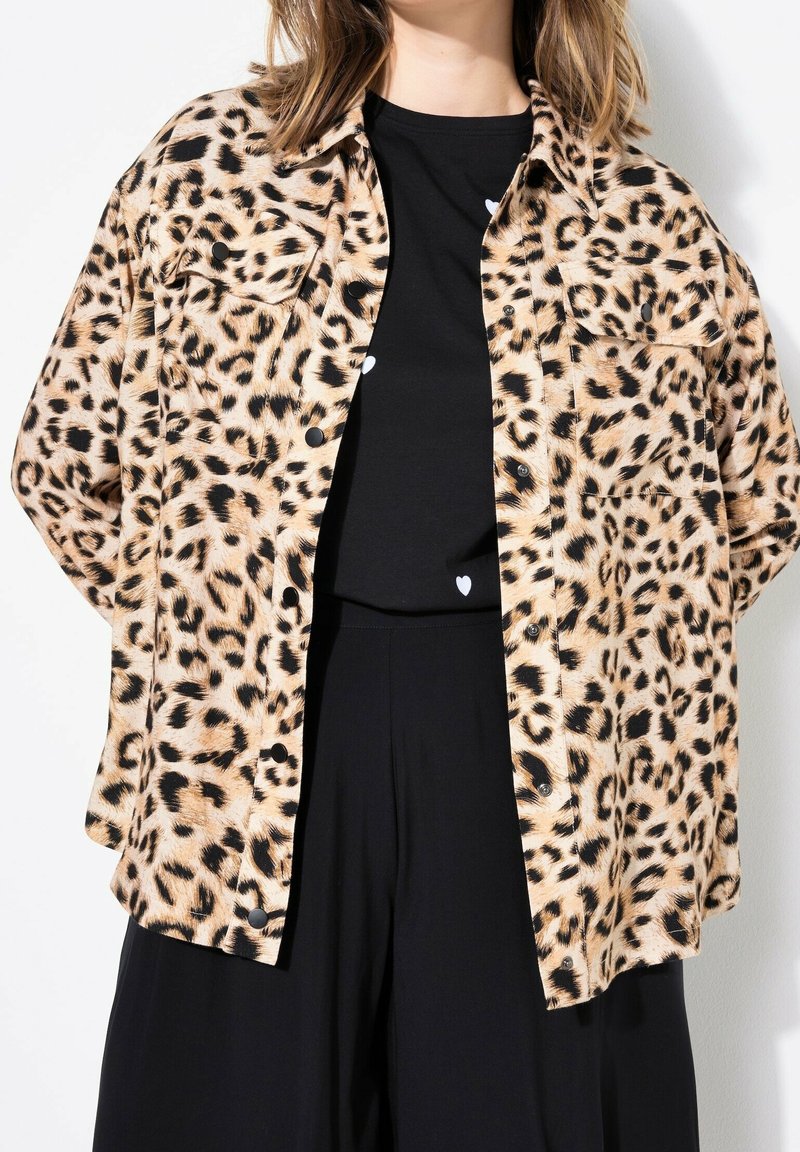 Leopard-print button-up shirt with a collar, short sleeves, and two front pockets, made of soft fabric; worn over a black top and pants.