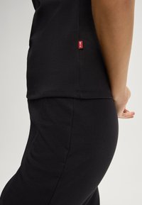 T-shirt noir en coton avec une fente sur le côté et une petite étiquette de marque rouge au bas. Sa texture lisse et son design ajusté améliorent le confort et la polyvalence.