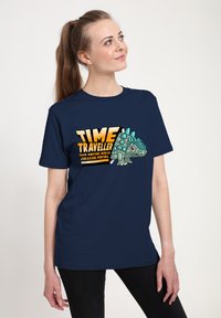 Námorně modré tričko vyrobené z bavlny, s grafickým designem dinosaura s ostny a textem, který říká "ČASOVÝ CESTOVATEL."