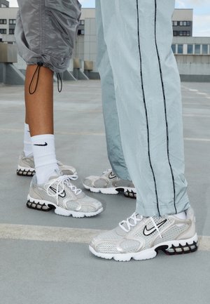 To personer iført lysegrå Nike-sneakers med hvide snørebånd og hvide Nike-sokker, står på en tagparkeringsplads.