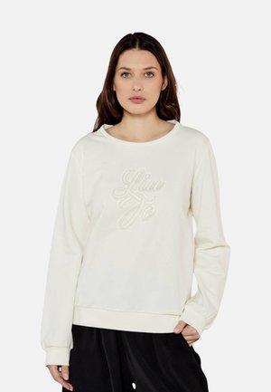 Vrouw draagt een wit sweatshirt met lange mouwen met "Liu Jo" in reliëf op de voorkant en een zwarte broek tegen een effen achtergrond.