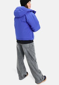 Lila Daunenjacke mit Kapuze, aus wasserabweisendem Material, kombiniert mit lockeren grauen Jeans und Schuhen mit brauner Sohle.