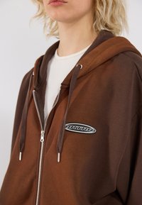 Brown zip-up hoodie with contrasting lighter brown sleeves, a soft texture, and a circular logo patch on the left chest area.