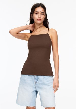 Top en coton marron ajusté avec de fines bretelles, associé à un short en denim clair avec un ourlet effiloché. Fond blanc uni.