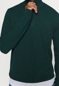 Mörkgrön pullovertröja med hög krage och halv dragkedja. Mjuk, strukturerad tyg med ribbade ärmslut och fåll, som har en lager-på-lager-look.