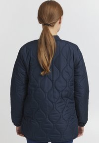 Marineblå quiltet jakke med en glat tekstur, der har en rundet krave og elastiske manchetter, vist bagfra med langt hår synligt.