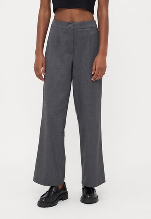 Persona con pantalones grises de talle alto y pierna ancha con botón y mocasines negros gruesos sobre un fondo liso.