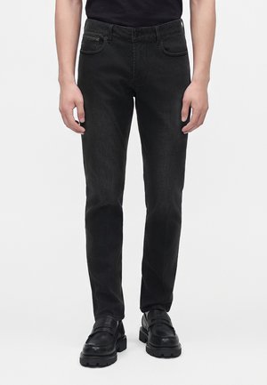 Persona con jeans negros ajustados y mocasines negros gruesos, de pie frente a un fondo claro y liso.