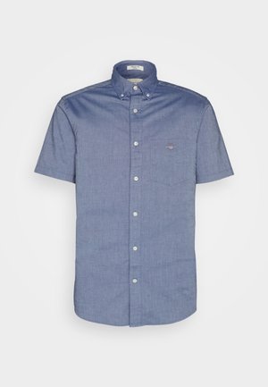 Camisa de manga corta abotonada en tela chambray azul claro, con un bolsillo en el pecho izquierdo que presenta un pequeño emblema de logo y un cuello estándar.