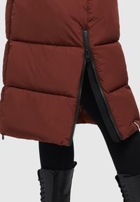 Hnedý puffrový vest s lesklým povrchom, ktorý má diagonálny zips na leme. Je semi-priehľadný a má vrstvený, prešívaný dizajn.